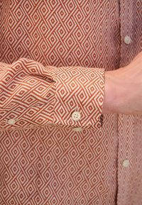 Camicia a maniche lunghe con motivo geometrico a diamante nei colori ruggine e crema. Polsini con bottoni color crema. Tessuto morbido al tatto.