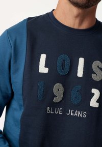 Sudadera color azul marino con letras bordadas texturizadas "LOIS 1962 BLUE JEANS." Presenta paneles laterales azules en contraste y un diseño de cuello redondo.