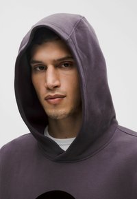 Sötét lila pamut kapucnis pulóver. Puha textúra, nagy fekete logóval. Fehér ing látható a kapucni alatt.