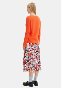 Orangener Strickpullover kombiniert mit einem plissierten, gemusterten Rock in Rot, Blau und Weiß; schwarze Plateauschuhe runden das Outfit ab.