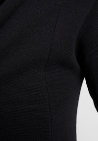 Mustaa neulottua kangasta teksturoidulla kuviolla, jossa on sileät saumat ja vartaloa myötäilevä muotoilu. Ei näkyviä metalliosia tai koristeita.