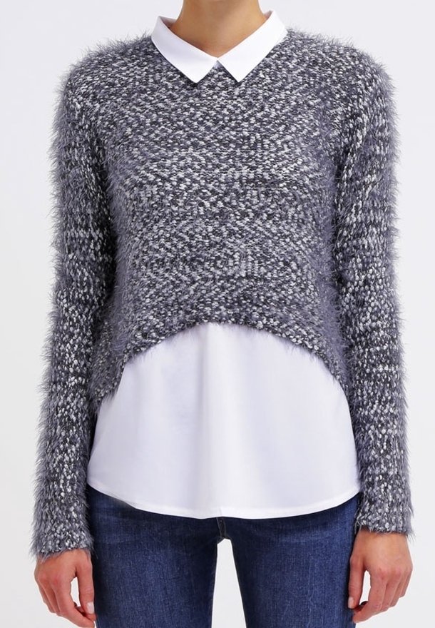 Personne portant un pull gris texturé et duveteux sur une chemise blanche à col et un jean bleu, montrant le haut du corps du cou jusqu'aux hanches.