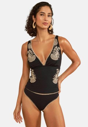 Woman wearing a black one-piece swimsuit with beige floral embroidery and hoop earrings, posing with one hand on her hip against a white background.