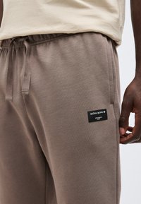 Κοντινό πλάνο ενός ατόμου που φοράει ελαστικό παντελόνι φόρμας ταμπά με ελαστική μέση και κορδόνι, με μια μαύρη ετικέτα της μάρκας Björn Borg στον μηρό.