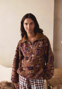 Brązowy polarowy sweter z zamkiem przy kołnierzu, z haftowanymi kwiatami w odcieniach fioletu i beżu. Noszony z w kratkę spodniami piżamowymi.