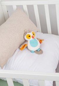 Jouet hibou en peluche aux grands yeux jaunes et ventre bleu, posé sur un oreiller blanc avec un coussin beige à l'intérieur d'un berceau blanc.