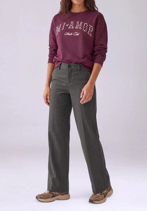 Un mannequin se tient dans un studio, vêtu d’un sweat-shirt bordeaux à motif graphique, d’un pantalon large gris anthracite et de baskets marron.