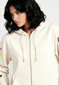 Kremasti duks na patentni zatvarač od mekog materijala, s vezicama za stezanje i dekorativnim vezom u različitim bojama na rukavima.