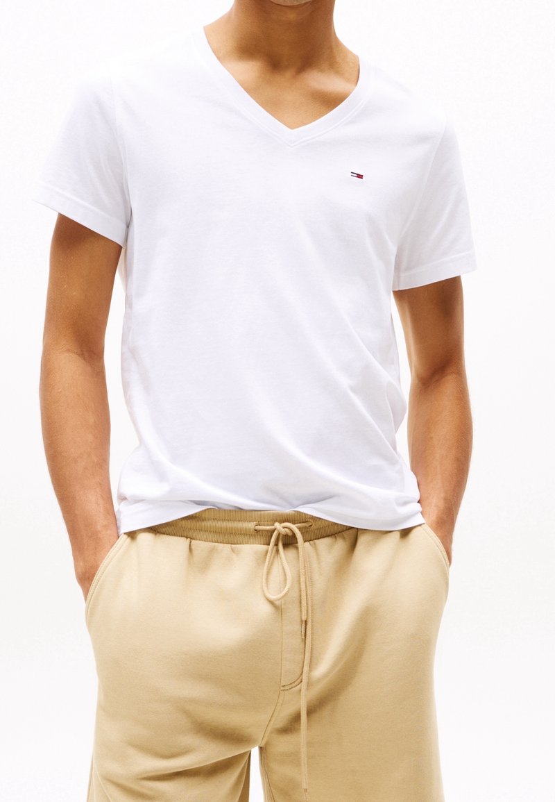 Hombre que lleva una camiseta blanca de cuello en V con un pequeño logo y pantalones beige con cordones, con las manos en los bolsillos.