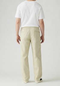 Homme portant un pantalon beige, un t-shirt blanc à manches courtes et des baskets noires et blanches, debout devant un fond clair uni.