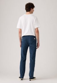 Bărbatul este întors cu fața, purtând un tricou alb, pantaloni slim-fit albaștri cu buzunare spate și adidași negri cu talpă albă.