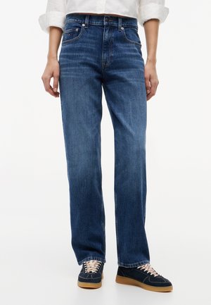 Personne portant un jean droit bleu foncé, une chemise blanche avec les manches retroussées, et des baskets bleu marine avec des semelles en gomme, debout sur fond blanc.