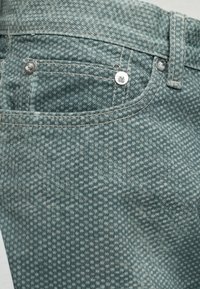 Gros plan de la poche arrière sur un jean en denim vert à motifs avec des rivets métalliques et une passants de ceinture au-dessus de la poche.