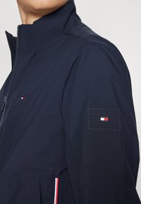 Giubbotto leggero blu navy con colletto alto, dotato di chiusura con cerniera e strisce di accento rosse, bianche e blu sui lati. Toppa con logo sulla manica.