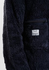 Pullover in pile blu navy con una superficie morbida e texturizzata; presenta una tasca con zip e un'etichetta in tessuto sul petto.