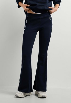 Persoon draagt marineblauwe flare joggingbroek met witte strepen aan de zijkanten en witte sneakers, tilt een donker sweatshirt iets op.