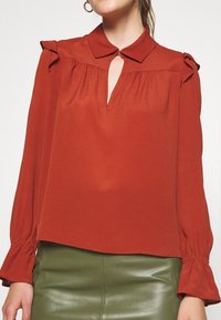Rostfarbene Bluse mit Kragen, Schlüsselloch-Ausschnitt und Rüschendetails an den Schultern. Lange Ärmel mit gerafften Manschetten, leichtes Material.