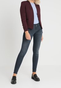 Blazer entallado burdeos sobre una camisa de rayas azul claro, combinado con jeans skinny oscuros y zapatos negros con cordones, creando una silueta ajustada.