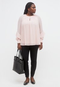 Γυναίκα που φοράει ανοιχτό ροζ μπλούζα με μακριά μανίκια και βολάν στις μανσέτες, μαύρο παντελόνι, μαύρα μπαλαρίνες και κρατά μια μεγάλη μαύρη τσάντα ώμου, κοιτάζοντας προς τα δεξιά.