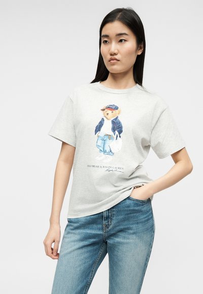 Νεαρή γυναίκα που φοράει ανοιχτό γκρι μπλουζάκι με μια αρκούδα σε στυλ casual και μπλε τζιν, ποζάροντας με το ένα χέρι στην τσέπη.