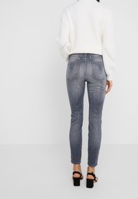 Jeans gris de corte slim, de talle medio, con bolsillos traseros, un leve desgaste y costuras de contraste, combinados con un suéter blanco de punto y tacones negros.