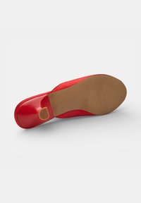 Zapato plano rojo de mujer mostrado desde el lado de la suela con suela de goma marrón y pequeño tacón antideslizante sobre fondo blanco.