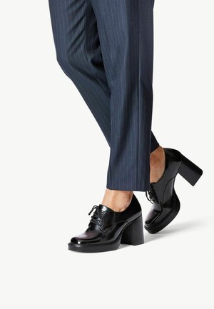 Czarne lakierowane oksfordy na obcasie z grubym, blokowym obcasem, z wiązaniem i gładką fakturą, w zestawieniu z spodniami w paski.
