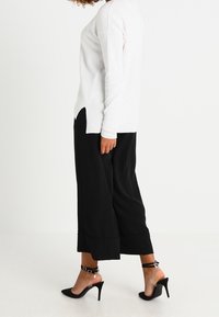 Top de manga larga blanco texturizado combinado con pantalones de pierna ancha negros. Zapatos negros de tacón alto con tiras en el tobillo completan el conjunto.