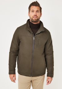 Olivgrüne Jacke mit durchgehendem Reißverschluss, hohem Kragen und zwei Seitentaschen. Strukturierter Stoff mit sichtbaren Nähten.