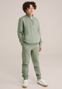 Svetlozeleni zip-up pulover s sprednjim žepom in ujemajočimi se cargo hlačami. Teksturna tkanina z rebrastimi manšetami in sproščenim fitom. Bele superge.