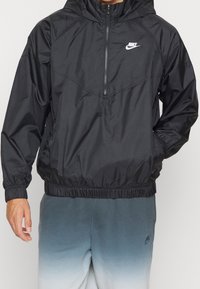 Musta kevyt tuulenpitävä takki, jossa on huppu, puolivetoketju edessä, joustavat hihansuut ja alahelma. Nike-logo rinnassa. Liukas kankaan rakenne.