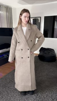 Μπεζ διπλόπατο παλτό με υφασμάτινη υφή, μεγάλα πέτα, μαύρα κουμπιά και πλαϊνές τσέπες, στιλισμένο πάνω σε λευκό τοπ και μαύρο παντελόνι.