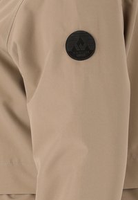 Beige jas met een gladde textuur en een ronde, zwarte patch met een bergenlogo op de mouw gestikt.