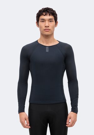 Un homme pose avec un maillot de cyclisme noir à manches longues et ajusté, ainsi qu’un short noir, sur un fond clair uni.