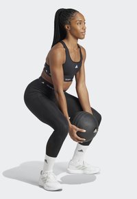 Svart atletisk crop-top og leggings med teksturert design, som holder en svart medisinball. Brukt med hvite sko og stripete atletiske sokker.