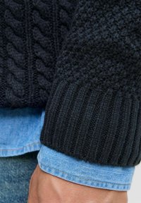 Pull en tricot noir avec motif en relief et poignets côtelés, superposé sur une chemise en denim bleu clair avec un poignet visible et un tissu texturé.