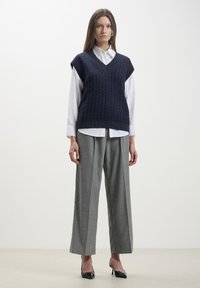 Gilet lavorato a maglia navy con scollo a V, caratterizzato da un motivo a cavo, indossato sopra una camicia bianca a maniche lunghe e abbinato a pantaloni grigi a gamba larga.