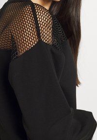 Sudadera negra con detalles de tul en los hombros, con mangas largas y un corte relajado, que resalta una mezcla de texturas.