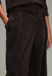 Ciemnobrązowe spodnie sztruksowe z pionowymi prążkami, wyposażone w boczne kieszenie i luźny fason. Tkanina ma miękką, teksturowaną powierzchnię.