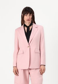 Blazer rosa con spalle strutturate, rever a punta e due tasche frontali. Presenta un bottone dorato ed è abbinato a una sciarpa nera.