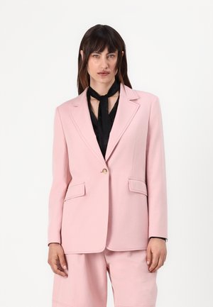 Rosa blazer med strukturerte skuldre, notched lapeller og to frontlommer. Har en gullknapp og er kombinert med et svart skjerf.
