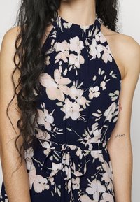 Granatowa sukienka w kwiaty z dekoltem halter, jasnoróżowymi wzorami kwiatowymi i wiązaniem w talii. Miękka, zwiewna tkanina o teksturowanym wyglądzie.