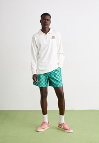 Hvitt langermet polo-skjorte med glidelås, grønn rutete shorts, hvite sokker og rosa Nike joggesko, stående på et grønt gulv.