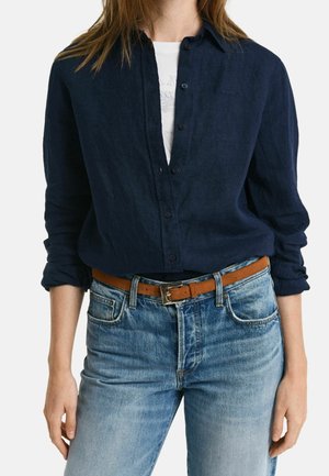 Personne portant une chemise bleu foncé à boutons sur un haut blanc, associée à un jean bleu clair et une ceinture en cuir marron.