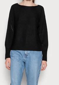 Schwarzer Strickpullover mit Schiffs-Ausschnitt, überschnittenen Schultern, gerippten Bündchen und offenen Strickdetails an den Ärmeln. Kombiniert mit hellblauen Jeans.