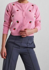 Kvinna iklädd en rosa knappad cardigan med röda jordgubbsmönster och hög midja mörkgrå plisserade byxor, poserande med en hand påMidjan.