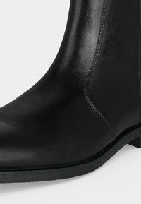 Gros plan d'une botte noire en cuir à la cheville montrant une texture lisse, des coutures latérales, un bout arrondi et un talon bas avec une semelle en caoutchouc.