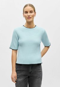 Pull en maille c ôtelée bleu clair à manches courtes, avec un bord festonné noir au niveau du col et des manches, associé à un jean gris foncé.