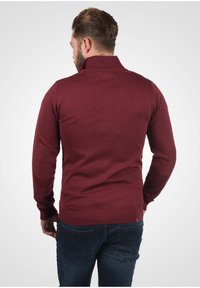 Burgundska pletena majica s visokim ovratnikom, rebrastim manžetama i donjim rubom, koja sadrži laktne zakrpe i uski kroj, uparen s tamnim trapericama.