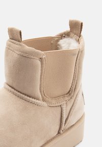 Bottine en suede beige avec une doublure intérieure douce, panneau latéral élastiqué, détails de couture et une petite languette à l'arrière.